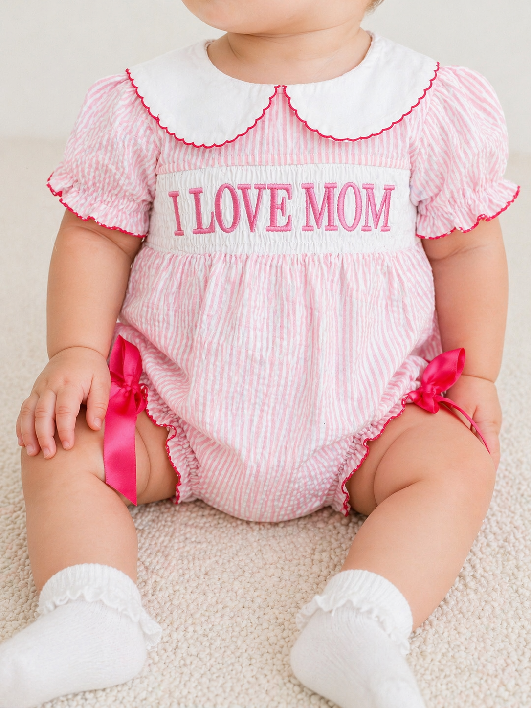 A baby sits on a light-colored carpet wearing the Tiny Tot Bundles 'I Love Mom Bubble'—a pink and white striped romper with puff sleeves, white collar, pink trim, and "I LOVE MOM" embroidery on the front.