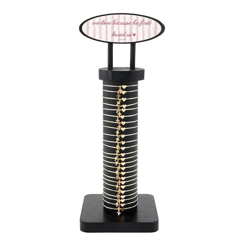 A Faithworks by Creative Brands “Heart Thread Bracelet Display - The Power of the Cross” holds multiple gold cross bracelets, featuring a top sign that reads “We love because he first loved us” with a small heart on a striped pink and white background.
