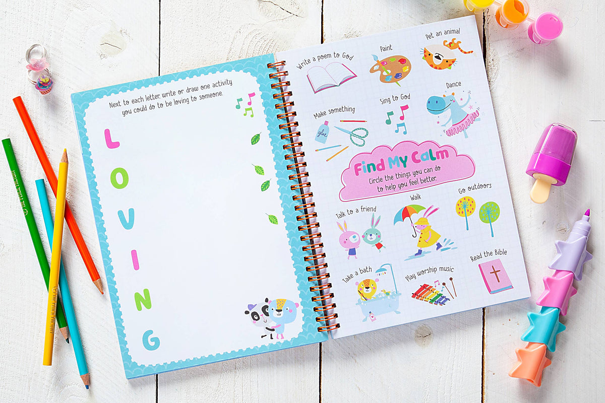 An open God Made Me Journal Book by BroadStreet Publishing Group, LLC is displayed on a white table alongside colored pencils and star-shaped crayons. The pages showcase a LOVING acrostic and a Find My Calm activity with playful, colorful illustrations.
