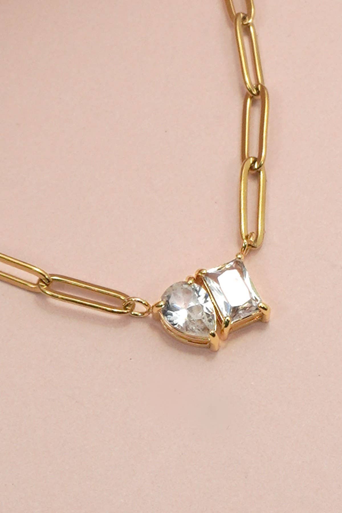 JOSSLYN by wall to wall’s Gold Rhin Necklace features elongated gold links and two side-by-side pendants—one round and one square clear gemstone—set in gold, shown against a pale pink background.