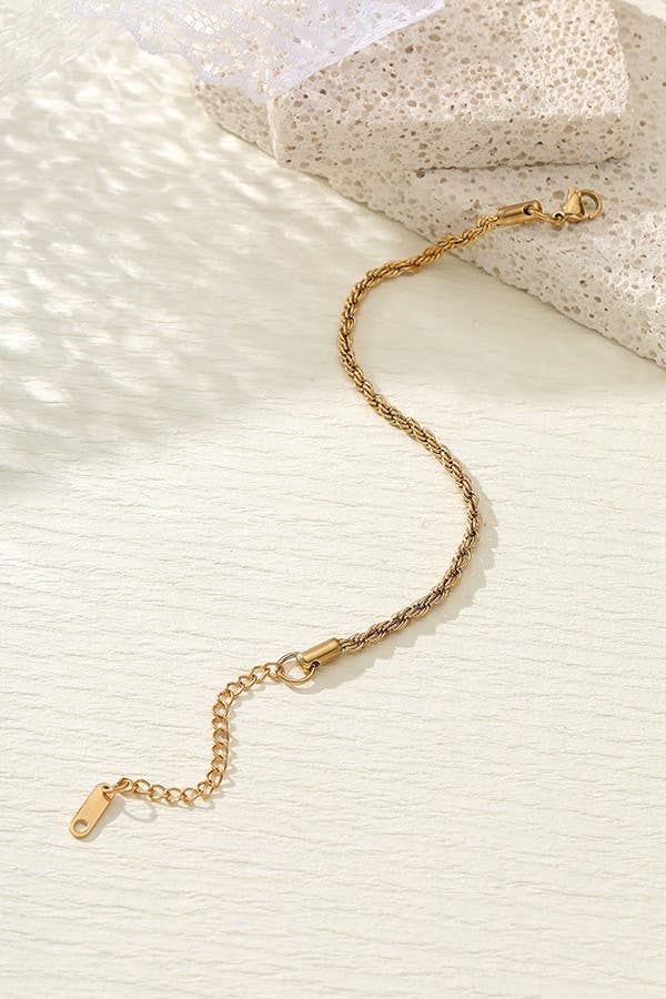A Dainty Gold Bracelet by JOSSLYN from wall to wall, featuring an adjustable clasp, rests on a textured light surface next to lace fabric and a stone-like object.
