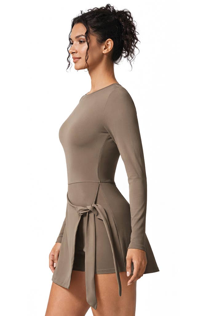 A woman stands in profile wearing the Color 5 Mocha Long Sleeve Athletic Dress, a fitted mini dress featuring a waist tie. Her curly hair is styled in a loose bun as she looks forward against a plain white background.