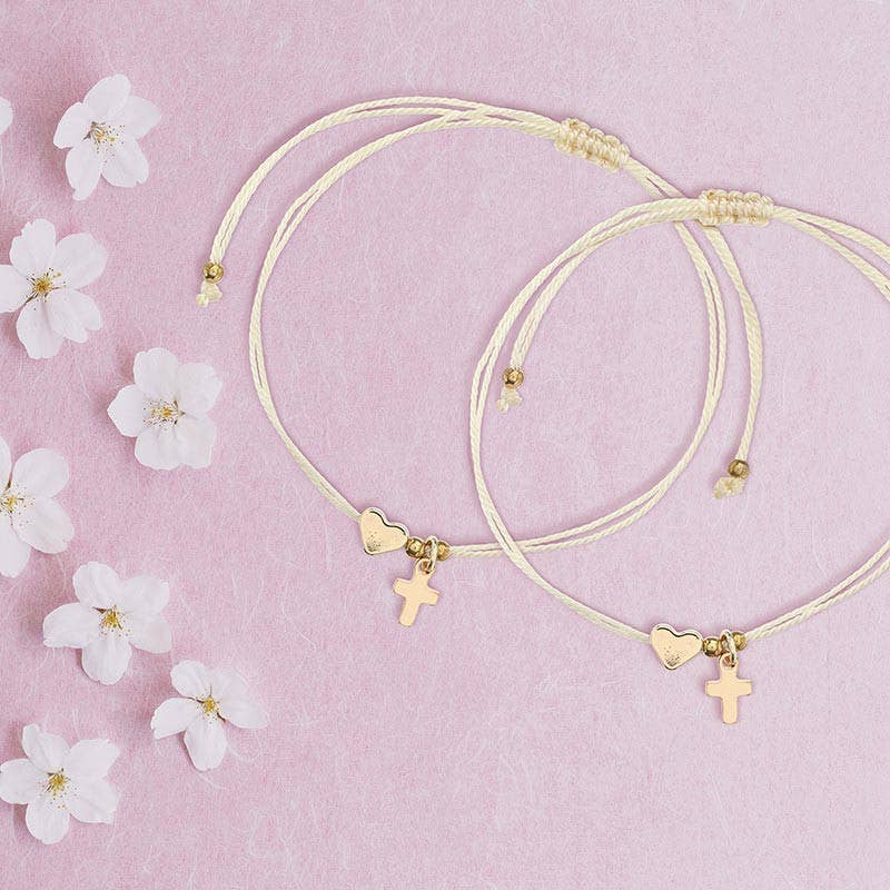 Two cream-colored Heart Thread Bracelets from Faithworks by Creative Brands’ “The Power of the Cross” collection, each featuring gold heart and cross charms, displayed on a pink background and surrounded by small white flowers.