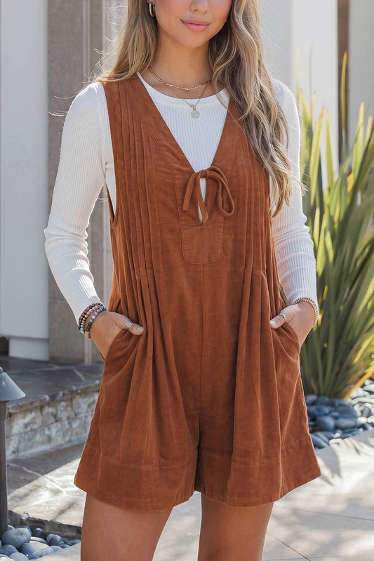 A woman stands outdoors near plants and a stone walkway, wearing a white long-sleeve top layered under the EVELA INC Corduroy Tie V-Neck Sleeveless Romper, which features front pockets.