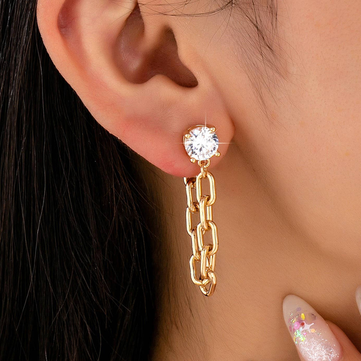 Close-up of an ear adorned with the JOSSLYN by wall to wall Stud Hoop and Drop Link Earrings, featuring a gold chain-link design and a large, round, clear gemstone stud. The person has long dark hair and glittery, decorated nails near their face.
