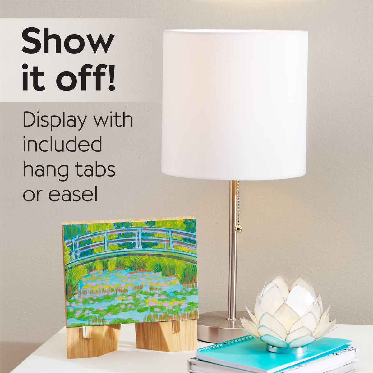 A small “Paint by Number Museum Series - The Japanese Footbridge” canvas by Faber-Castell / Creativity for Kids is displayed on a wooden easel next to a modern white lamp and a lotus-shaped decoration on a desk, with text inspiring you to showcase your art and paint like the Masters.