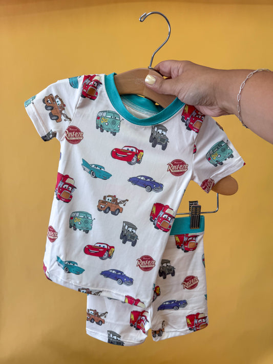 A hand holds up a hanger displaying the Tiny Tot Bundles Route 66 Dreams Lounge Set—an adorable toddler matching shirt and shorts set with colorful cartoon cars on a cheerful yellow background.