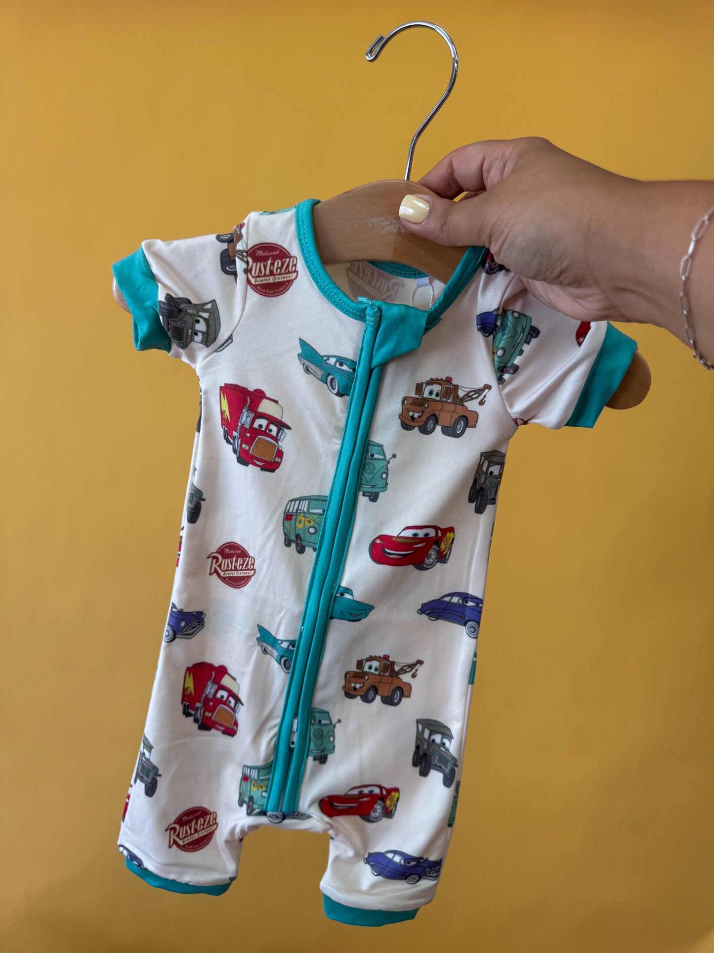 A hand holds up the Tiny Tot Bundles Route 66 Dreams Short Zippy, featuring a Cars-inspired print and teal trim, against a solid yellow background.