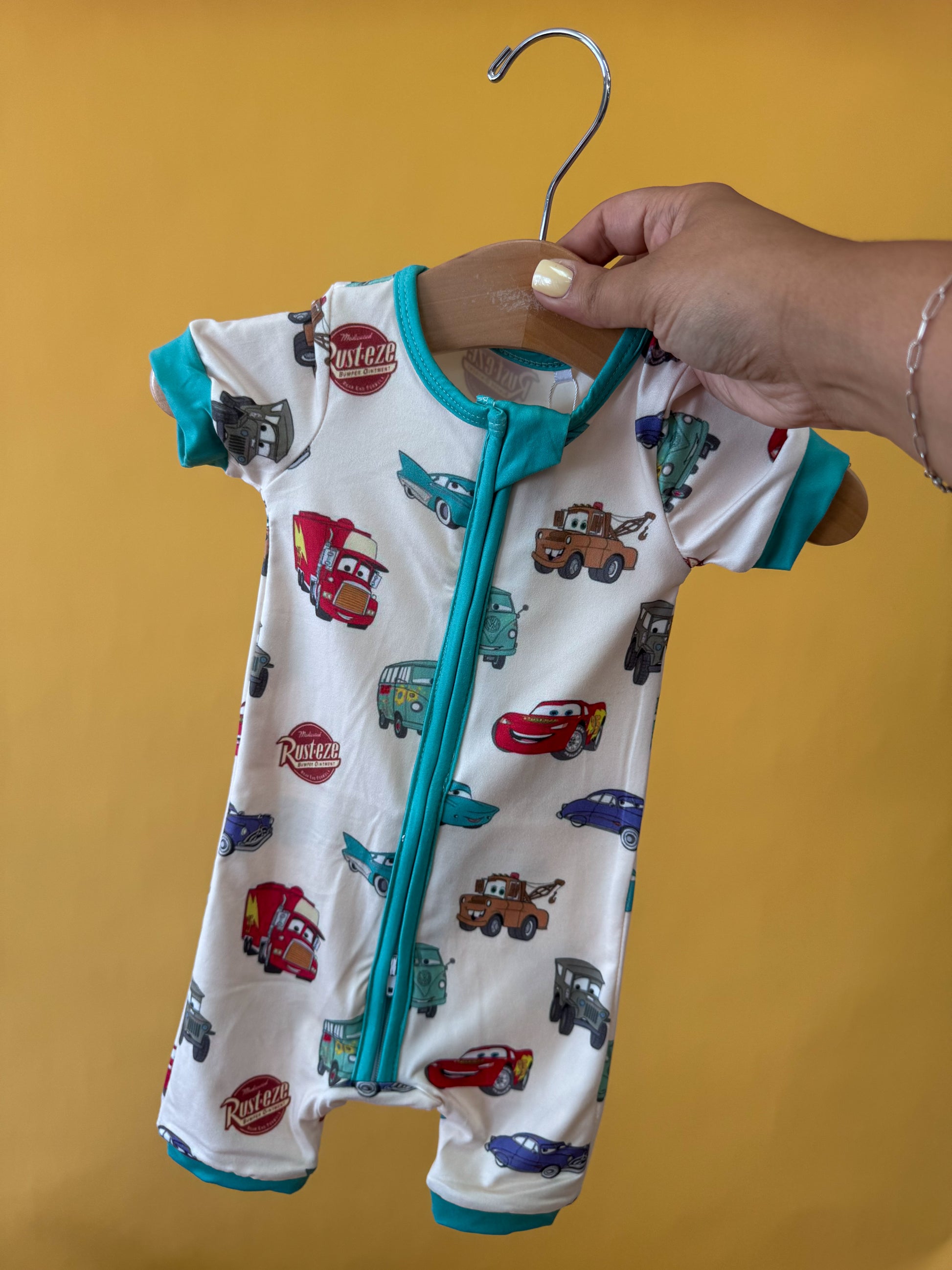 A hand holds up the Tiny Tot Bundles Route 66 Dreams Short Zippy, featuring a Cars-inspired print and teal trim, against a solid yellow background.