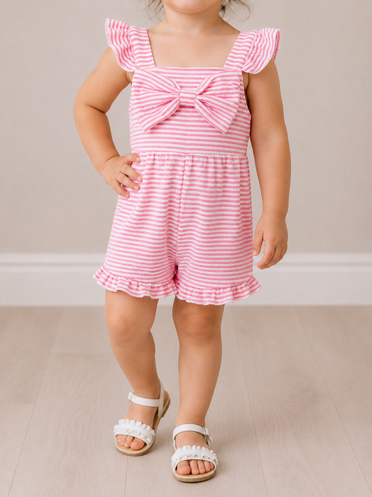 A young child in the Tiny Tot Bundles Sugar Stripe Bow Romper, featuring pink and white stripes, ruffled sleeves, and a large front bow, poses with one hand on their hip and wears white sandals on a light wooden floor.