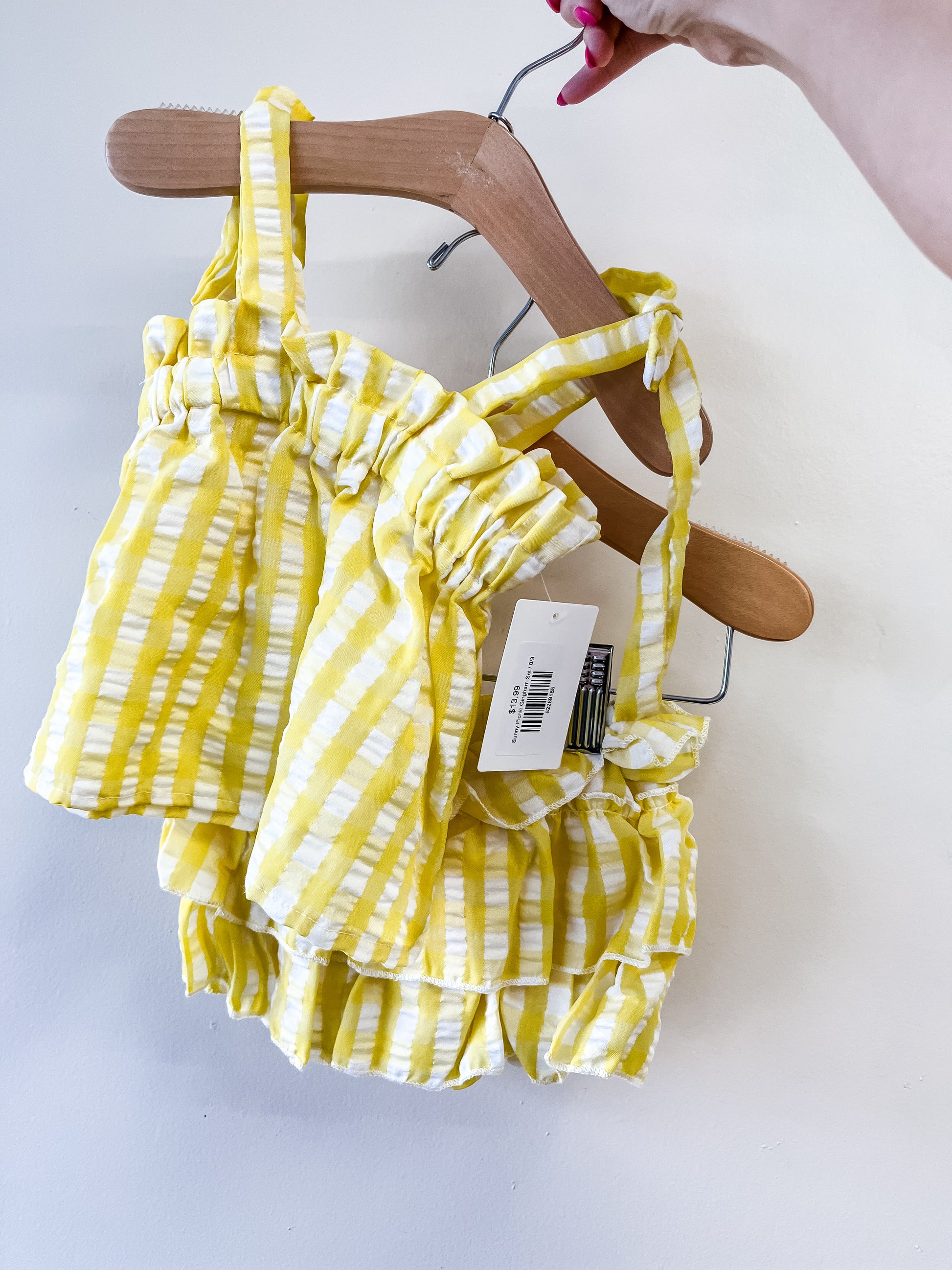 A hand holds a hanger with the Tiny Tot Bundles "Sunny Picnic Gingham Set," a yellow and white striped ruffled baby outfit with tags attached, shown against a plain white background.