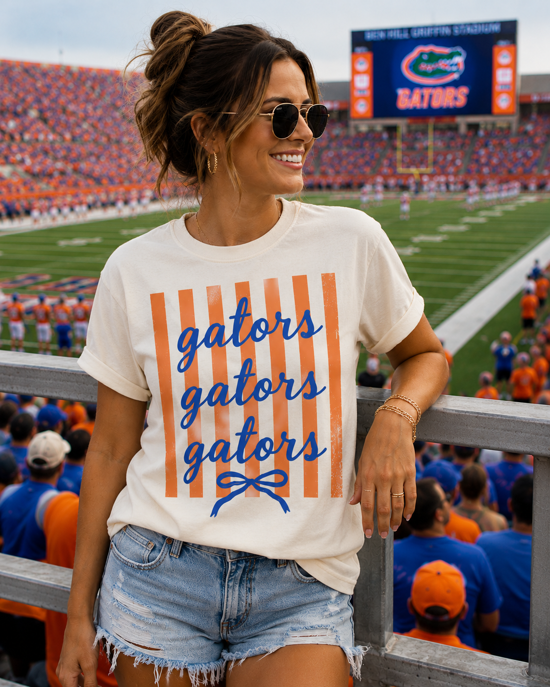 A woman in sunglasses and a Three Threads Branding Co. Gators Stripe Graphic t-shirt stands smiling at a football stadium, with fans and the field in the background.