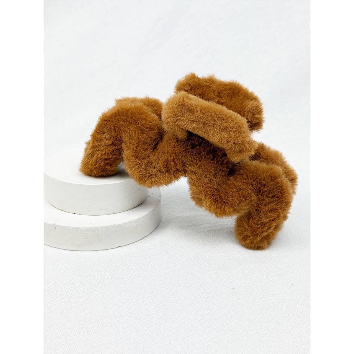 A Jumbo Fur Wavy Hair Claw Clip by Love and Repeat, featuring a fuzzy brown arch design, rests on two round white platforms against a plain white background.