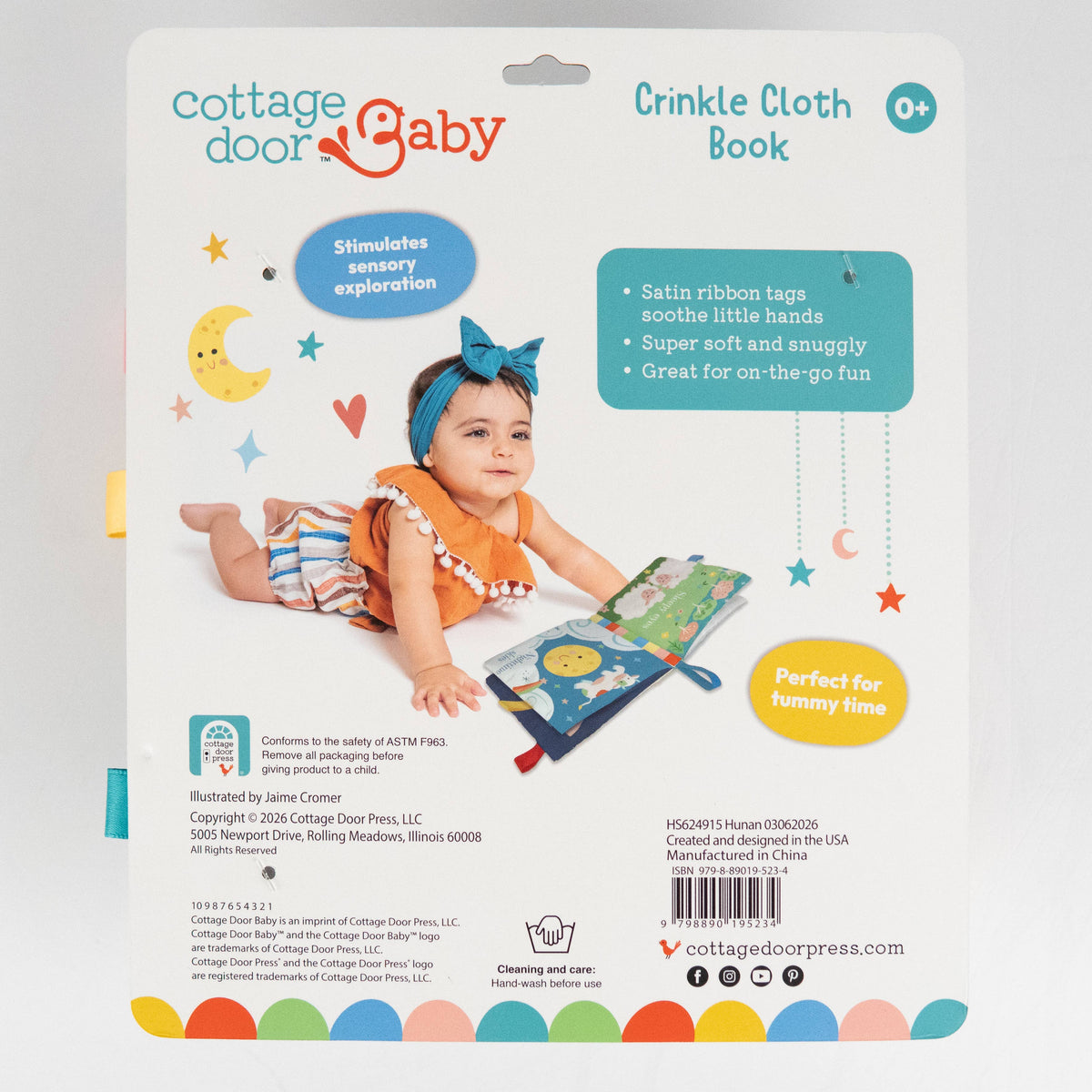 A baby in a blue headband lies on her stomach playing with the colorful Sweet Dreams Cloth Book by Cottage Door Press. The packaging highlights features such as satin ribbons, sensory play, and tummy time suitability, making this baby sensory cloth book perfect for your little one’s daily routine.