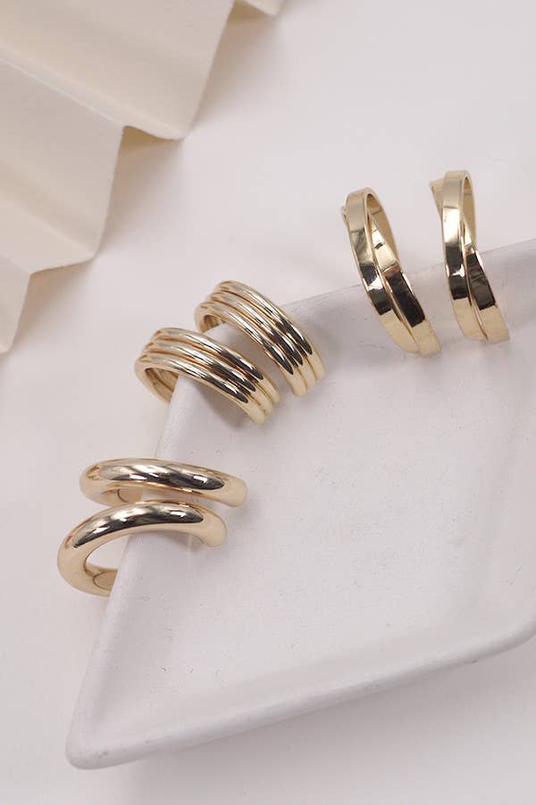 The Multi Hoop Trio Earring Set by JOSSLYN by wall to wall, featuring four pairs of shiny gold hoop earrings in varying thicknesses, is displayed on a white triangular ceramic dish and a white surface.