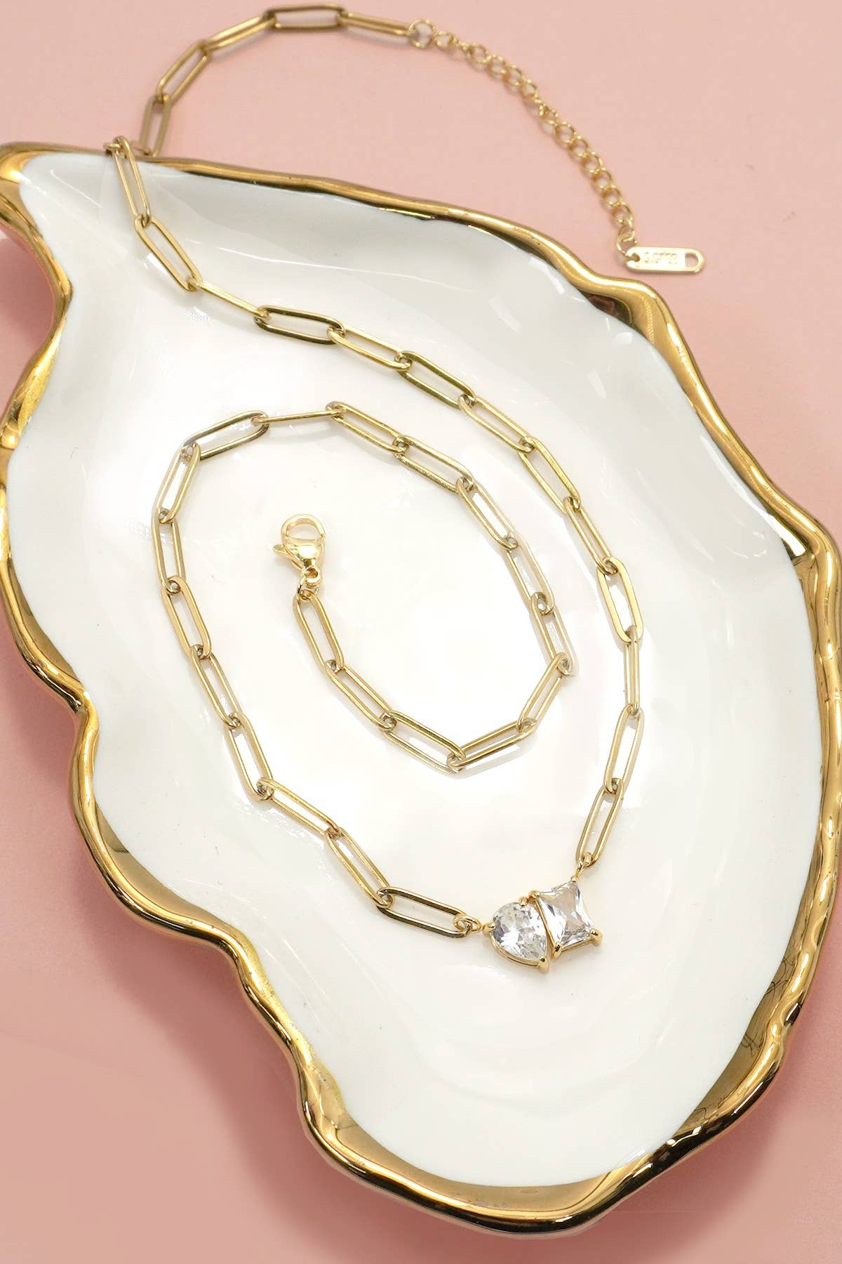 JOSSLYN by wall to wall’s Gold Rhin Necklace, featuring rectangular links and a teardrop-shaped gem, is elegantly displayed on a white dish with gold trim against a pink background.