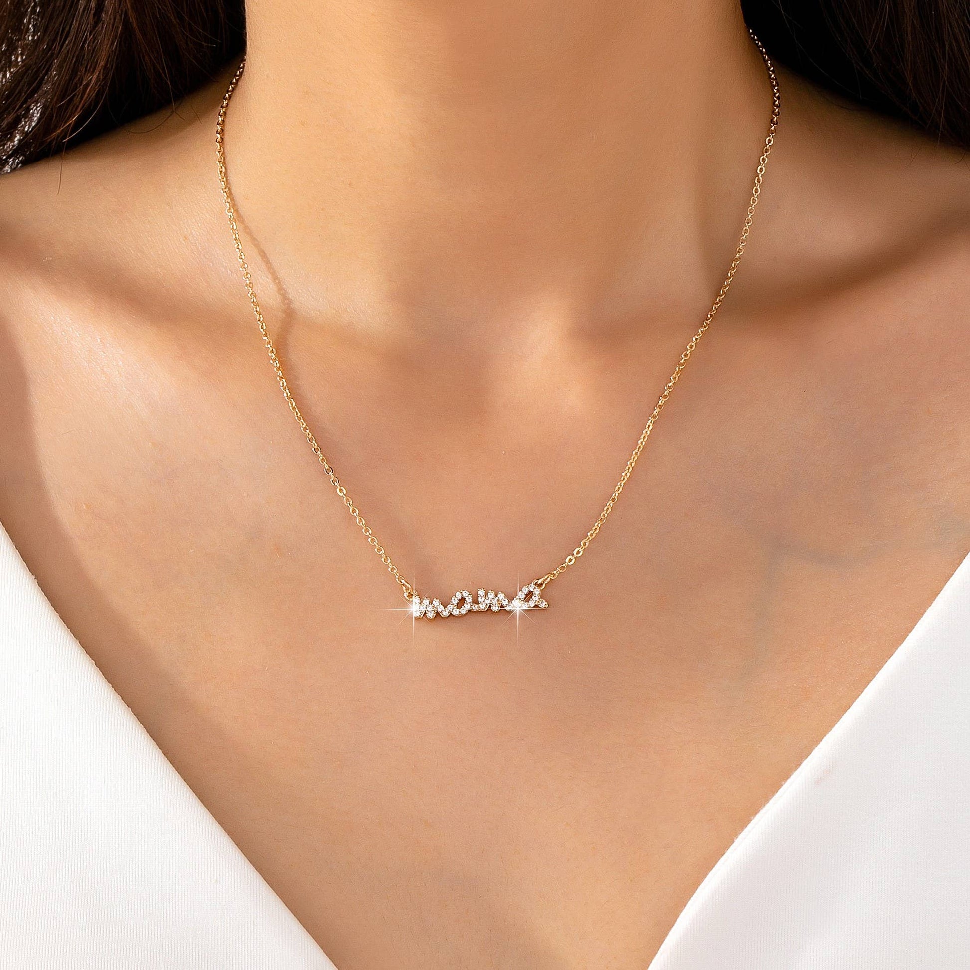A close-up of a woman’s neck wearing the Gold MAMA Necklace by JOSSLYN by wall to wall, featuring cursive lettering adorned with sparkling stones, paired with a white V-neck top.