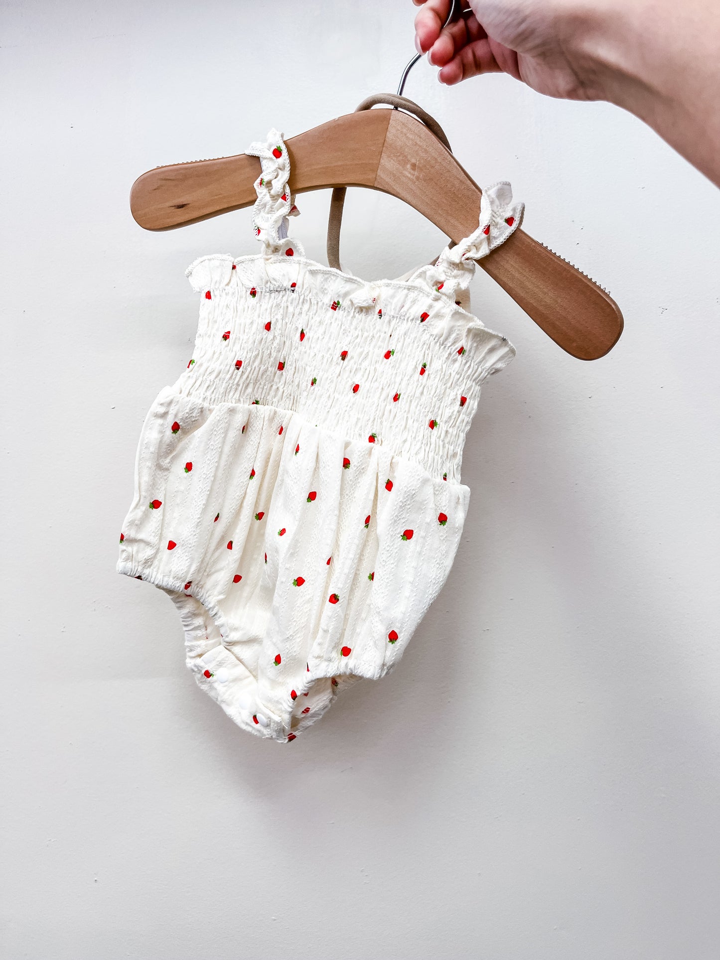 A hand holds the Tiny Tot Bundles Linen Strawberry Romper, a white baby outfit with red polka dots and ruffled straps, displayed on a wooden hanger against a plain white background.