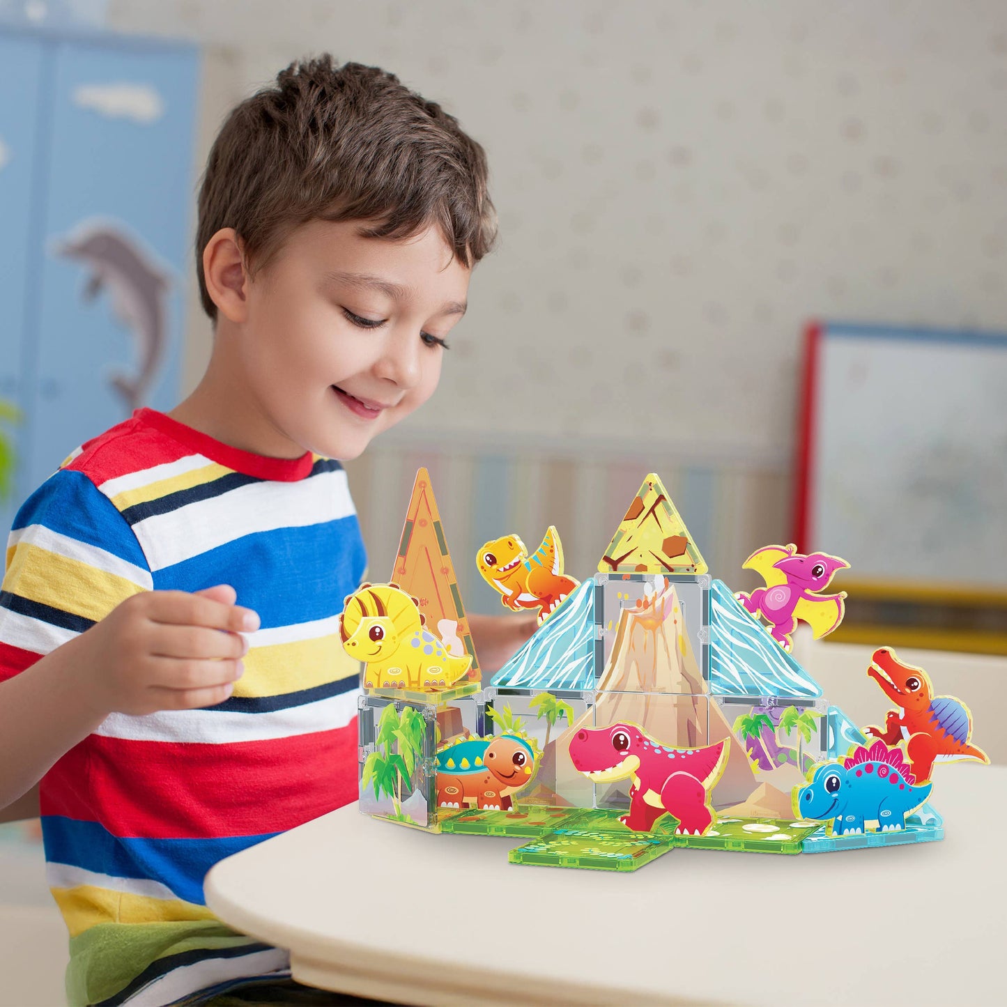 A young boy in a striped shirt smiles while playing with the PicassoTiles 26pc Dino Theme Magnetic Tile Building Set by PicassoTiles. Bright dinosaur figures and transparent magnetic tiles are arranged on the table before him.