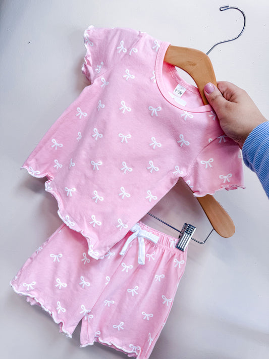 A hand holds the Little Miss Bows Cotton Set by Tiny Tot Bundles—pink short-sleeve top and matching shorts with small white bow patterns and ruffled edges, shown against a plain light background.