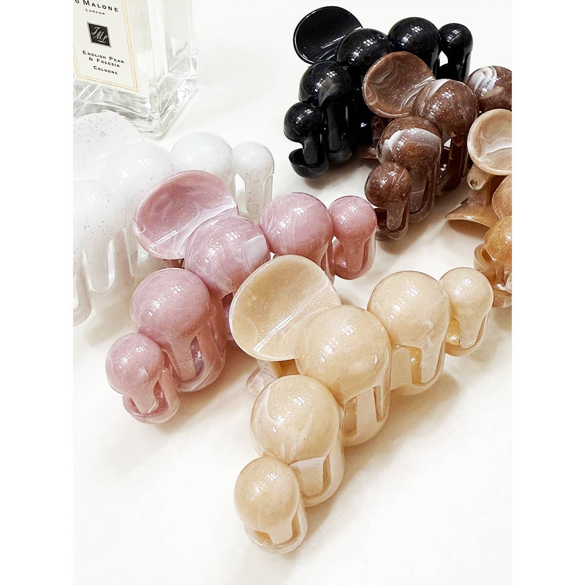 A selection of Love and Repeat Large Marbled Hair Claws, featuring matte finishes in beige, pink, white, brown, and black shades, displayed on a white surface beside a clear perfume bottle.
