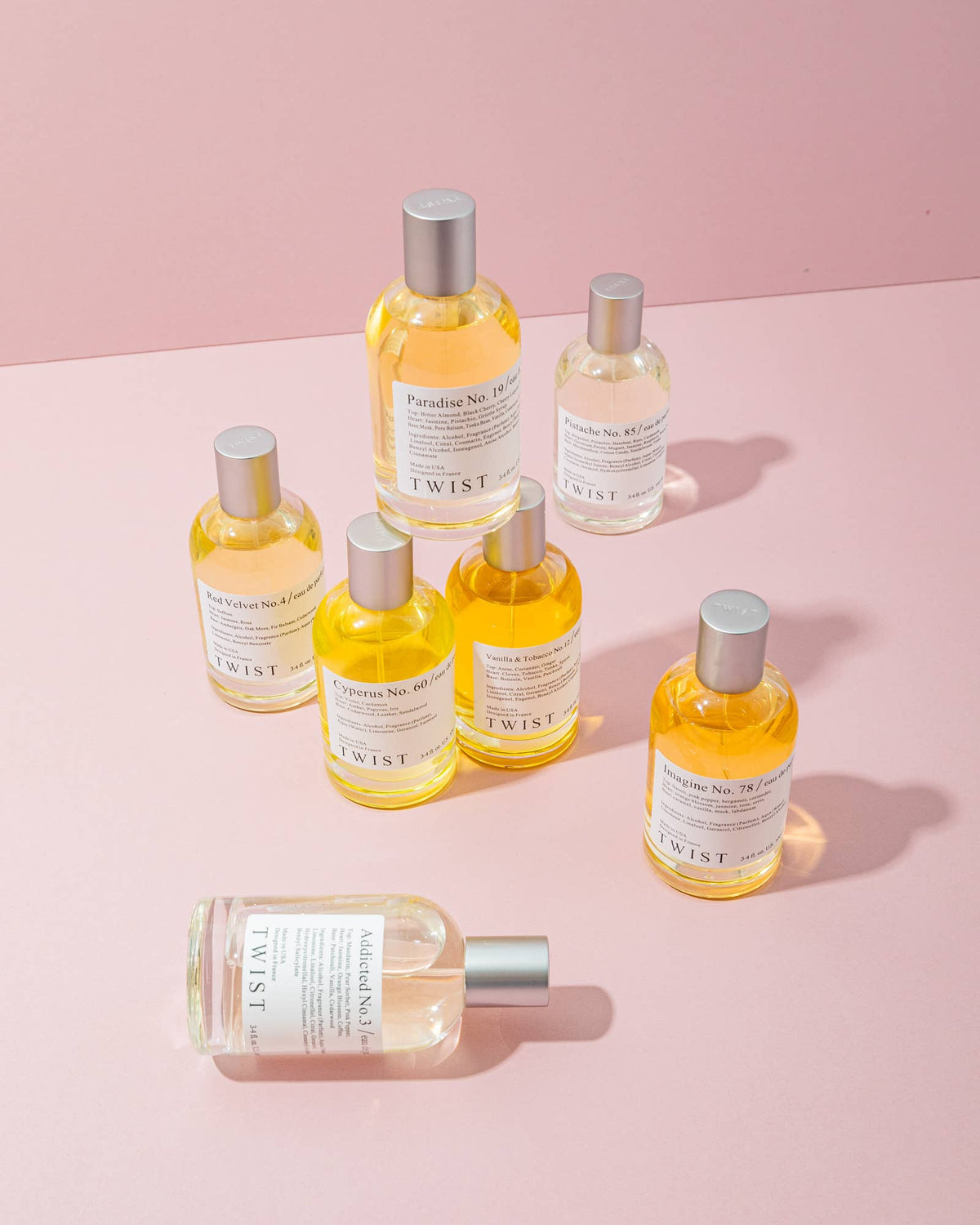 Seven bottles of House of Twist Sun Kissed No. 115 Perfume with silver caps are arranged on a pink surface, casting soft shadows. The bottles contain yellow and clear liquids—each offering a unique summer scent—and feature white labels with black text, making them an ideal long-lasting perfume for women.