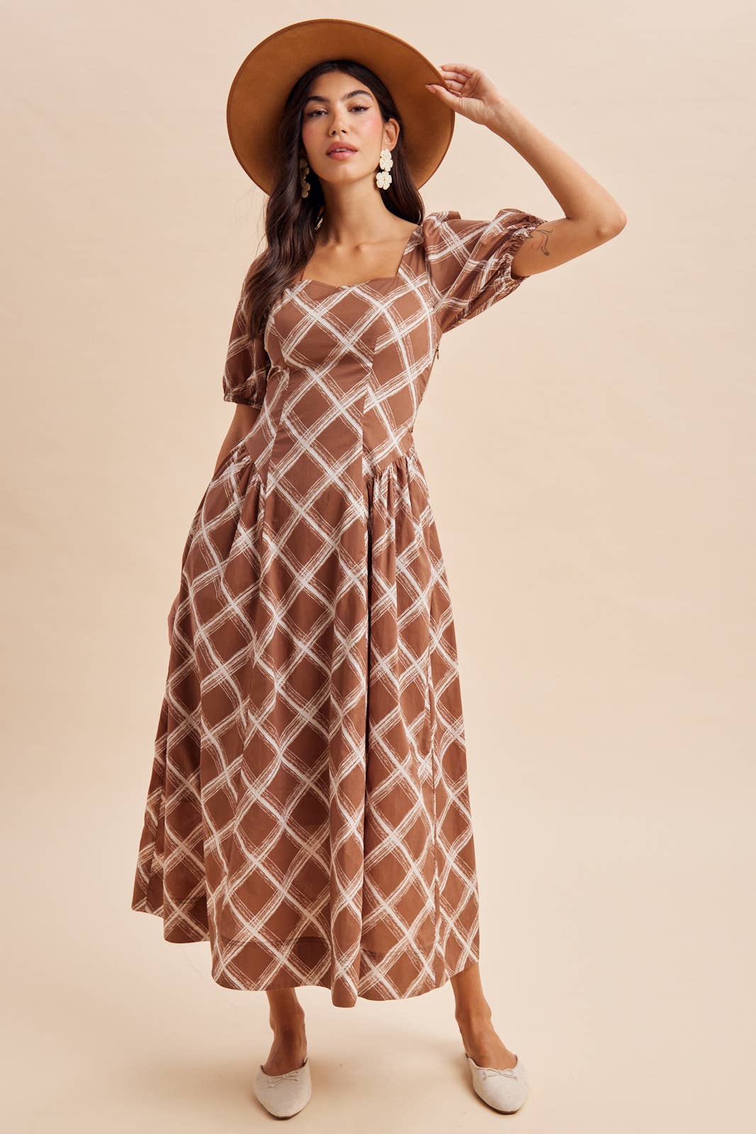 A woman wears an In Loom Plaid Square Neck Maxi Dress in brown, featuring a square neckline and puff sleeves, paired with a wide-brimmed hat and ballet flats.