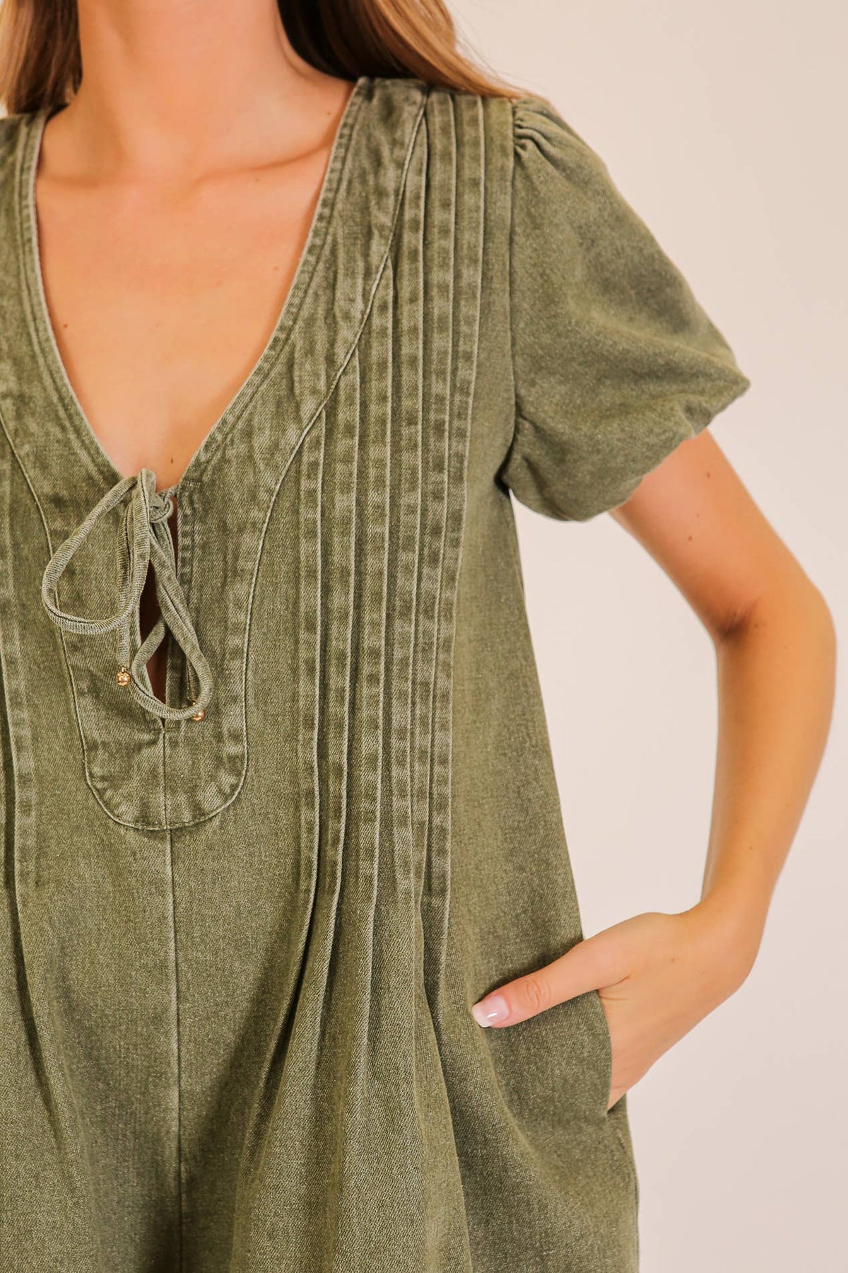 A woman wears the Lumiere Desert Muse Utility Romper—a short-sleeve, olive green one-piece featuring a deep V-neckline, front tie detail, vertical pleats, and convenient pockets. Only her torso and part of her left arm are visible.