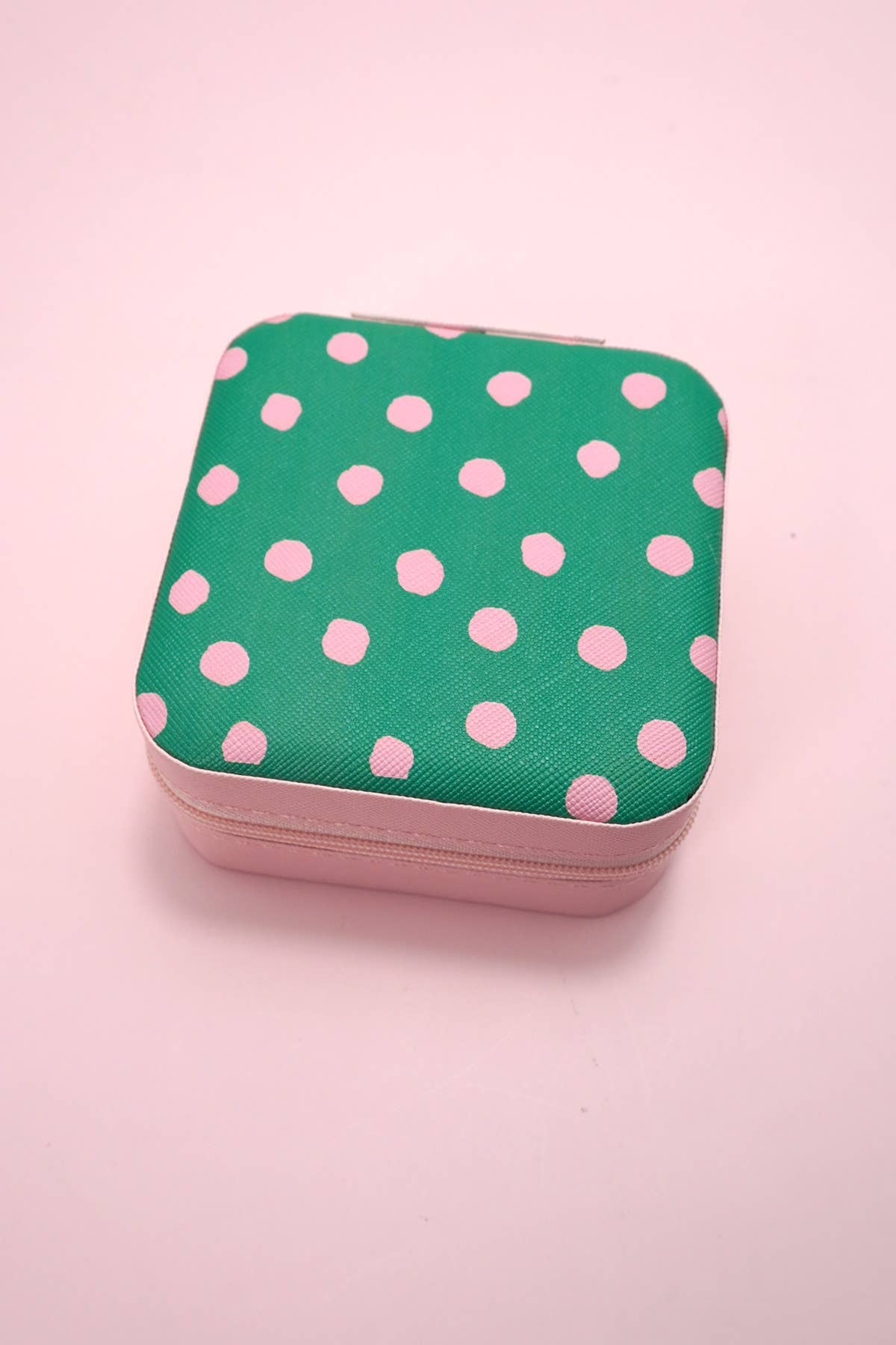 A compact, square Jewelry Organizer Travel Box Case by JOSSLYN from wall to wall, featuring a green lid adorned with pink polka dots, is displayed on a light pink surface.