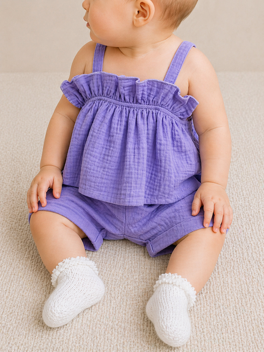 A baby in the Tiny Tot Bundles Lavender Breeze Short Set sits on a light carpet, gazing to the side. The outfit includes a sleeveless lavender top and matching shorts, paired with white knit socks featuring small decorative details.