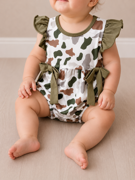 A baby sits on a light wooden floor wearing the Tiny Tot Bundles Green Camo Bow Romper, a sleeveless outfit featuring green, brown, and white camouflage, ruffled sleeves, and green waist bows. The baby's face is not fully visible.
