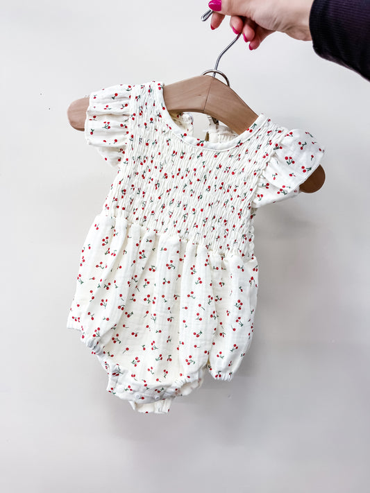 A hand holds Tiny Tot Bundles’ Cherry Sweetheart Bubble, a white baby romper with short ruffled sleeves, a smocked bodice, and tiny red cherry prints. The wooden hanger and the plain white background highlight its charming design.