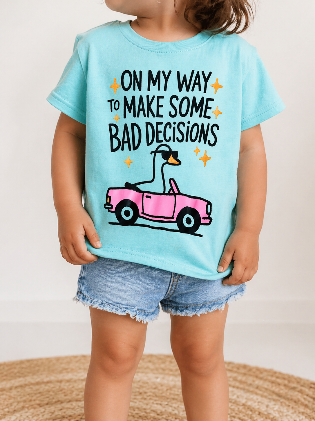 A young child wears the Wild Threads Co Bad Decisions t-Shirt—a light blue tee with a cartoon swan driving a pink car and the text "ON MY WAY TO MAKE SOME BAD DECISIONS"—paired with denim shorts.
