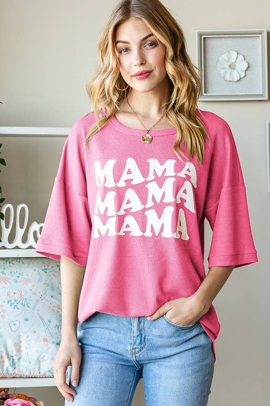 A woman stands indoors with one hand in her jeans pocket, smiling slightly at the camera while wearing the HEIMISH Solid Ribbed MAMA Shirt—a pink top featuring “MAMA” printed four times in bold white letters.
