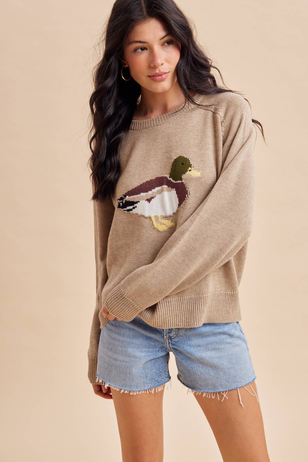 A woman wears a tan crew-neck In Loom Mallard Duck Knitted Sweater featuring a knitted mallard duck design on the front.