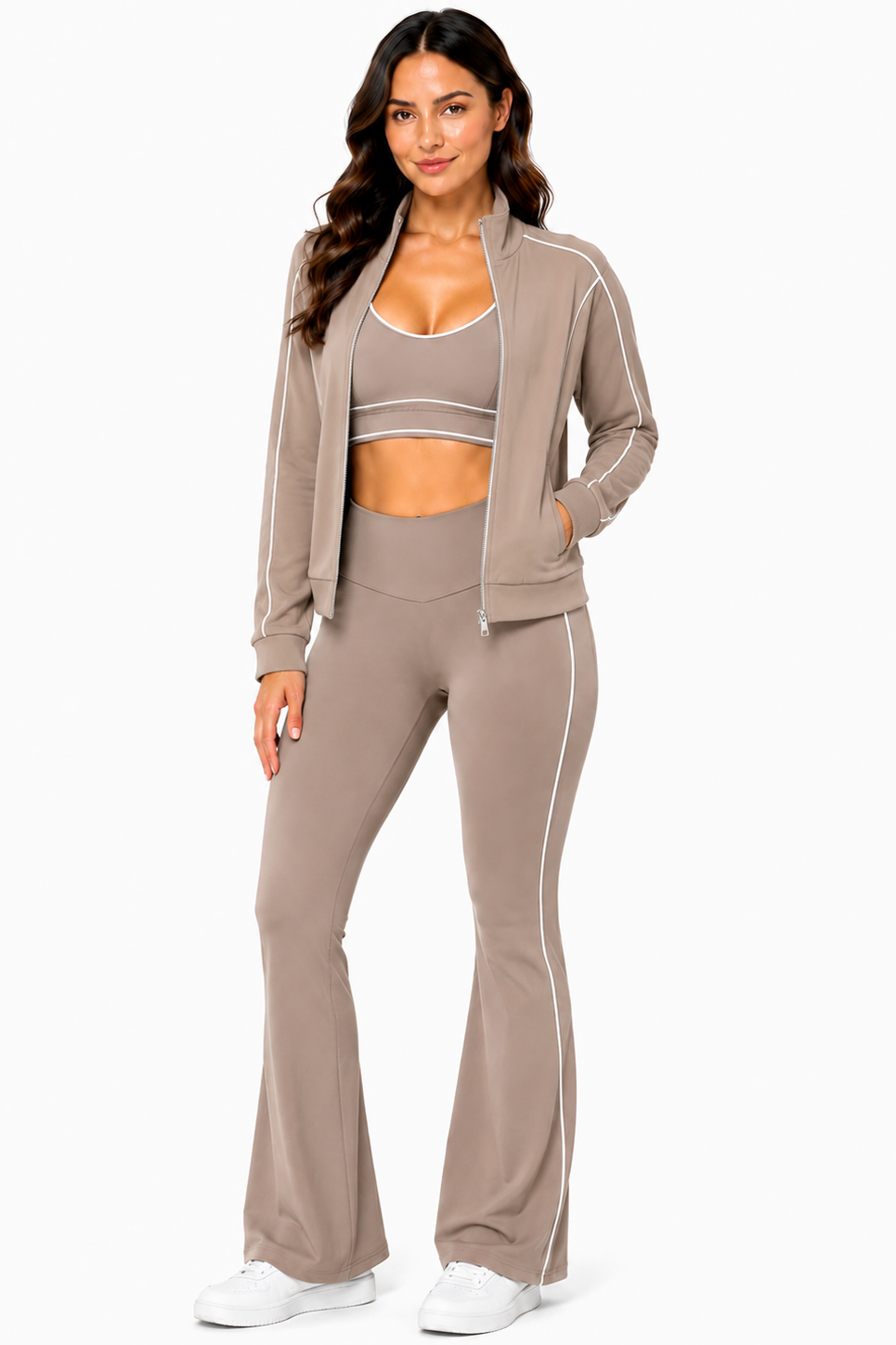A woman stands and smiles against a white background, wearing the Color 5 Mocha Muse 3-Piece Athletic Set—a taupe zip-up jacket, sports bra, and flared pants with white piping—paired with white sneakers.