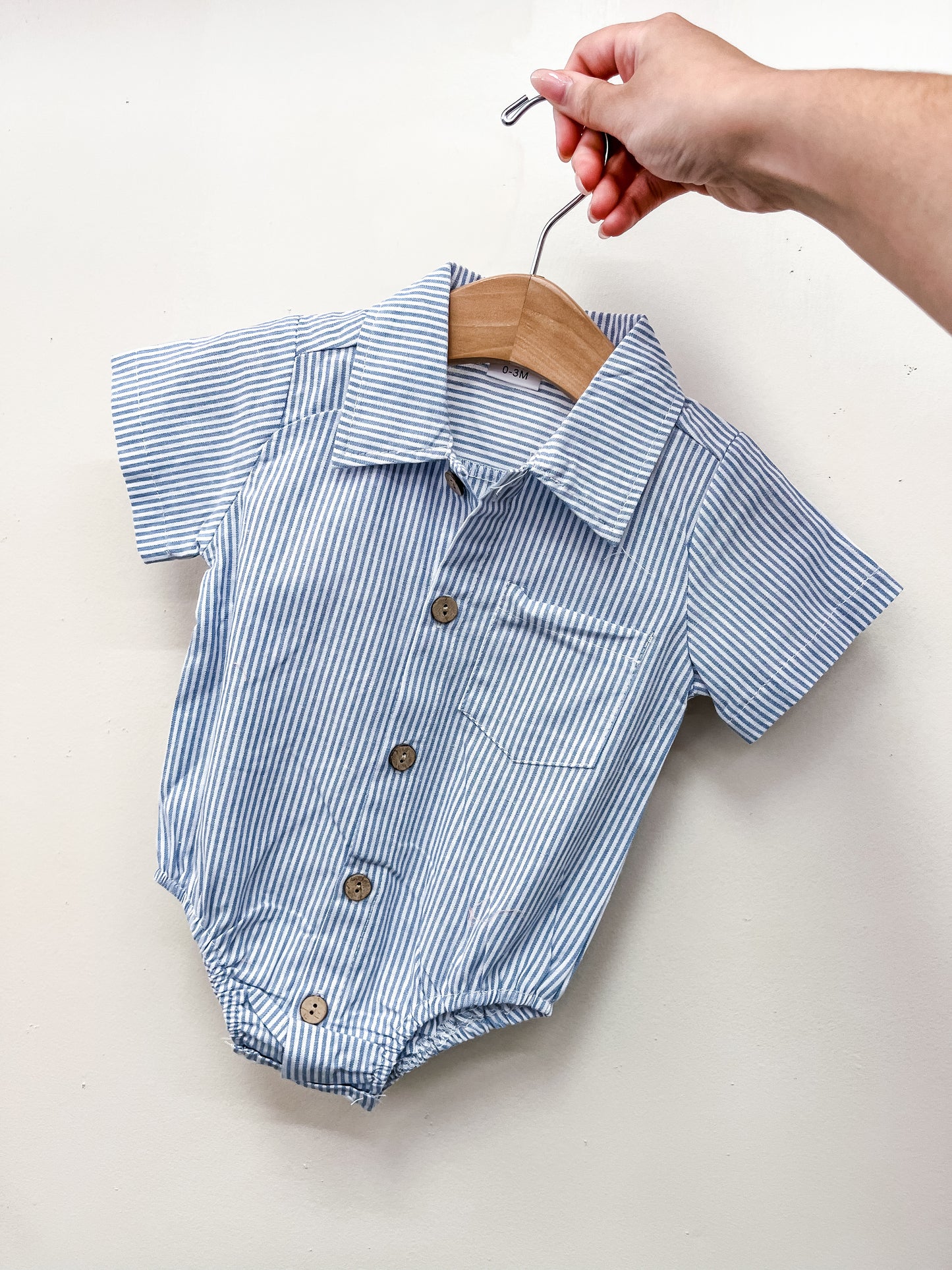 A hand displays the Tiny Tot Bundles Blue Stripe Collared Bubble—a baby blue and white striped short-sleeve romper with brown buttons and a small chest pocket—on a wooden hanger against a plain white background.