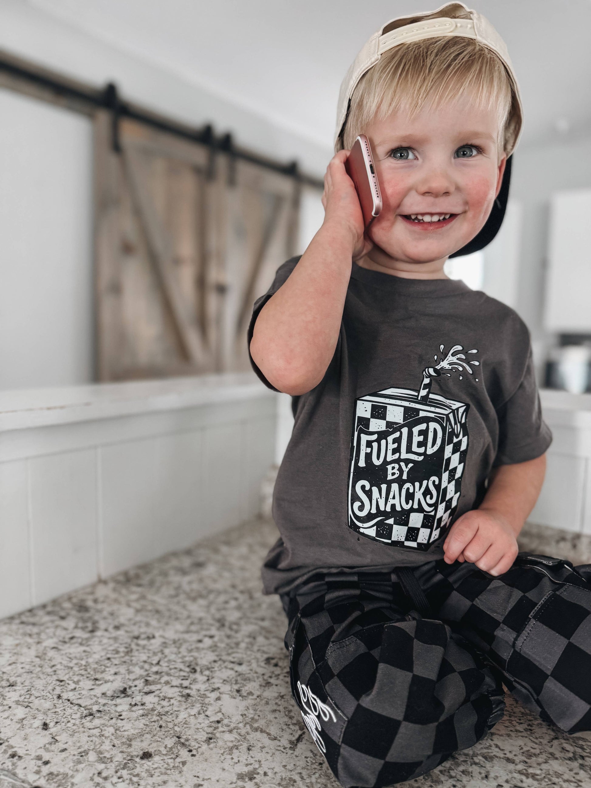 A smiling young boy in a backwards cap sits on a counter, holding a phone to his ear. He wears the Raising Brave “Fueled By Snacks” Toddler Tee featuring a checkered design, paired with matching checkered pants.