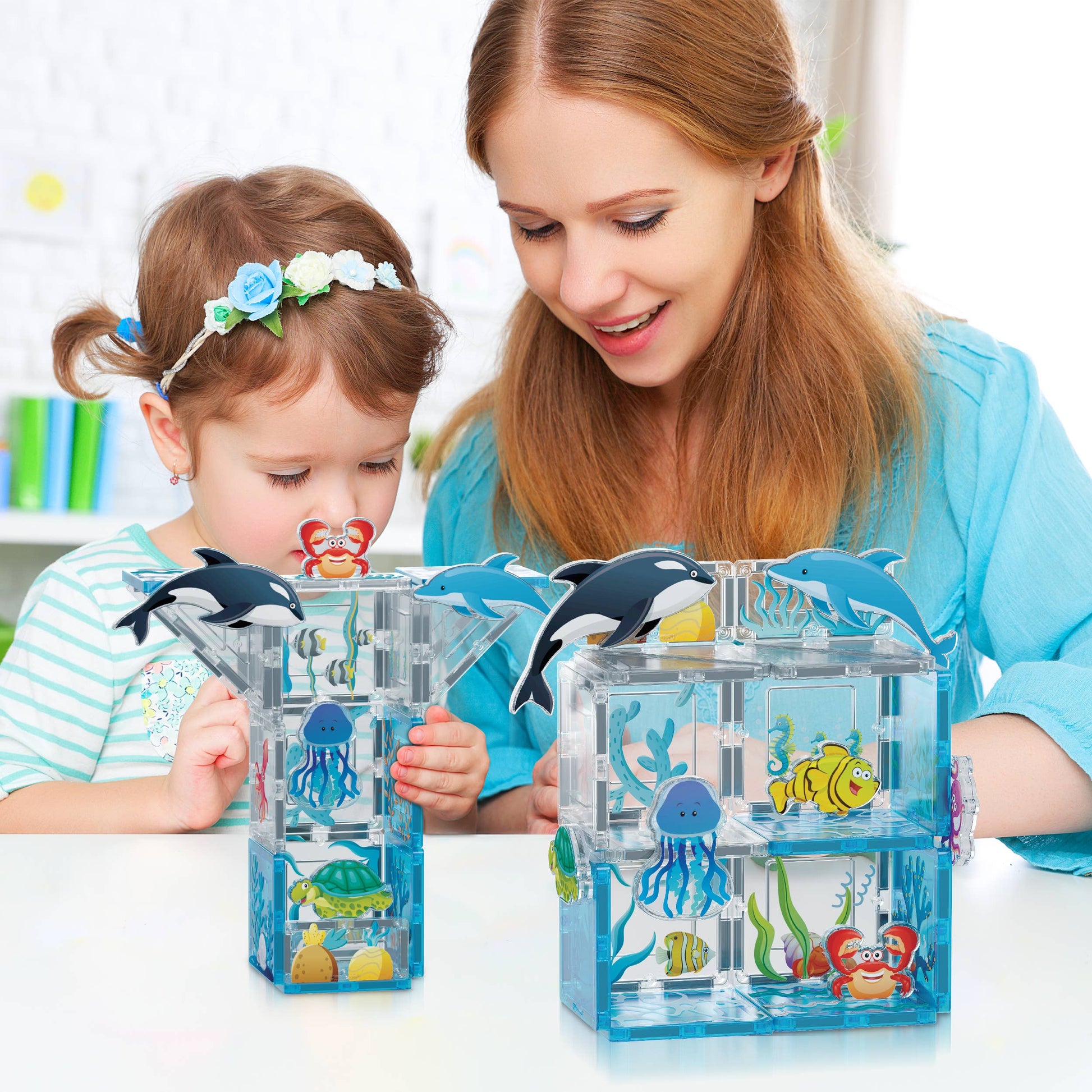 A woman and a child happily play with the PicassoTiles 26 Piece Marine World Magnetic Building Set, using clear plastic aquariums decorated with toy sea animals like fish, jellyfish, orcas, and crabs on a white table.