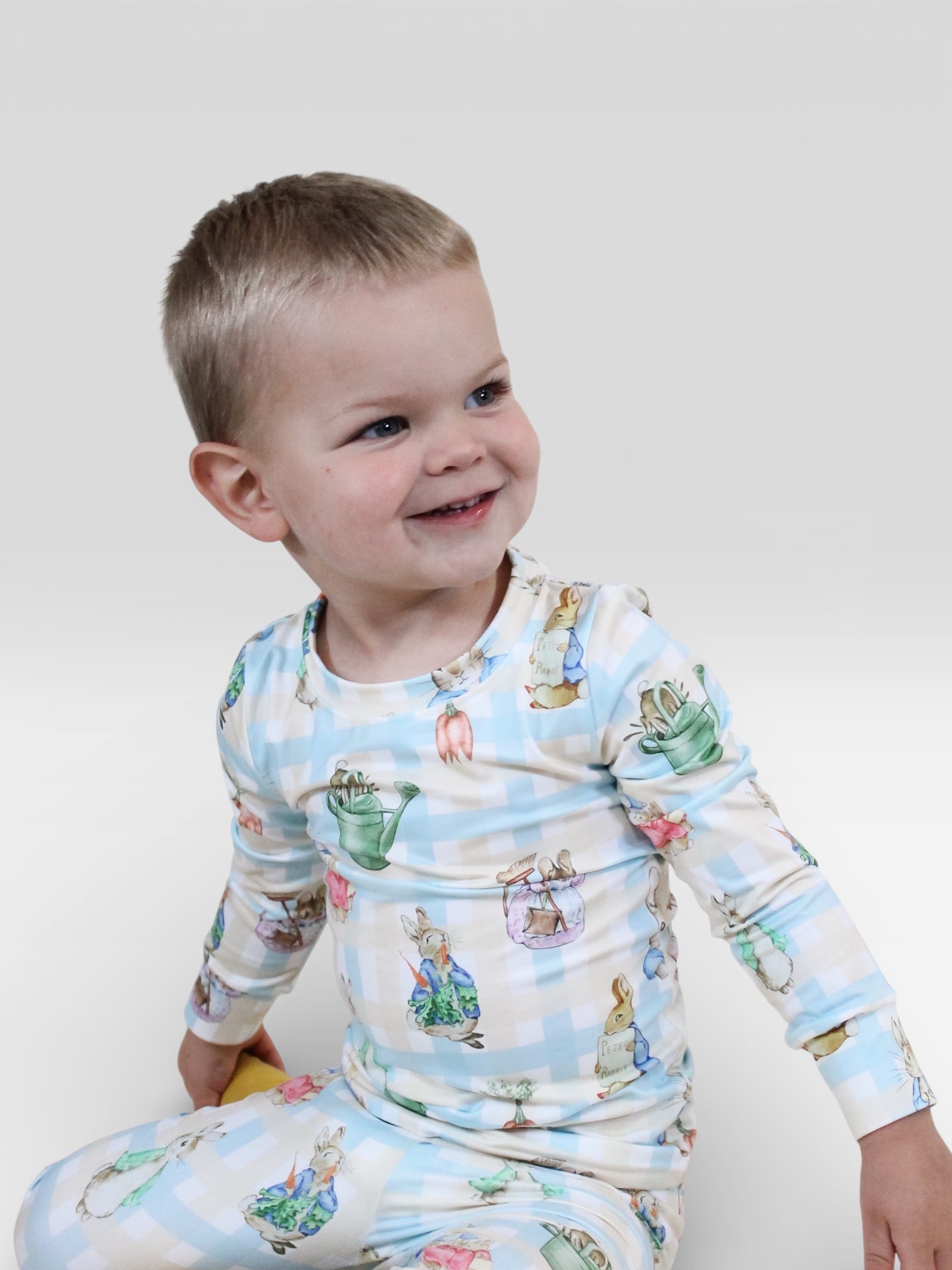 A young child with short blond hair smiles while wearing Tiny Tot Bundles' Peter's Easter Picnic Jammies Set, featuring light-colored checks with animal and teapot prints, seated against a plain white background.