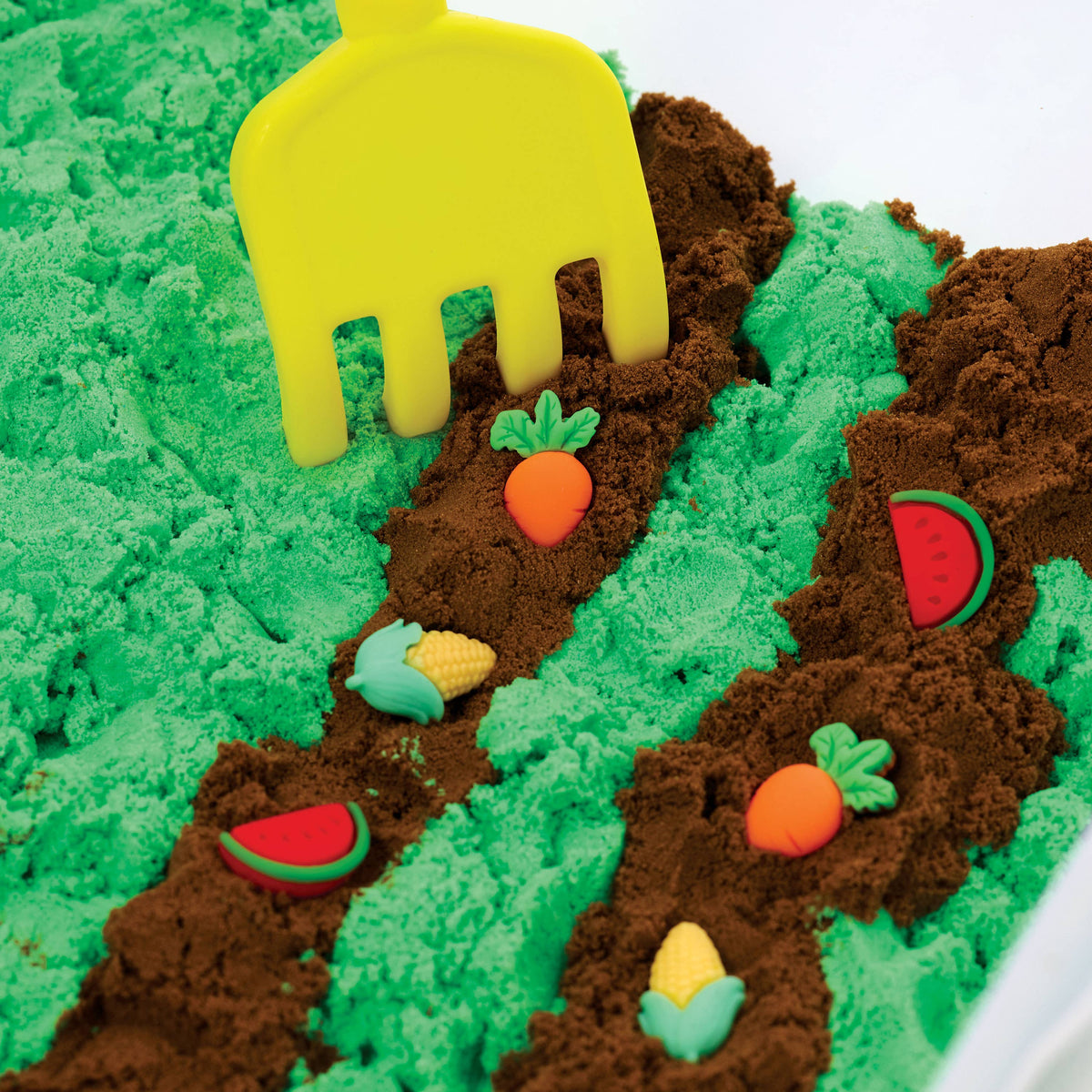 The Sensory Bin Farm Activity Kit for Kids by Faber-Castell / Creativity for Kids features a yellow toy rake nestled in green and brown sensory sand, alongside small toy fruits and vegetables such as carrots, watermelon slices, and corn arranged in neat rows. This farm-themed sensory bin encourages children to develop their fine-motor skills through hands-on play and exploration.