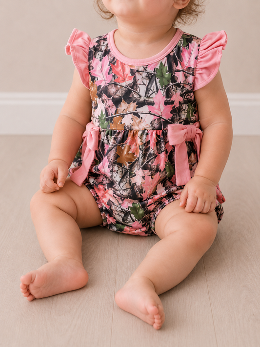 A baby, partially out of frame, sits on a light wooden floor wearing the Tiny Tot Bundles Pink Camo Bow Romper with ruffled sleeves and pink bows.