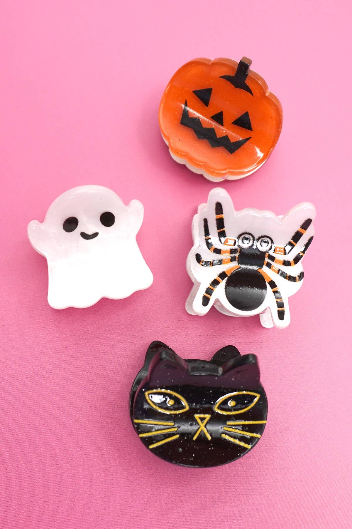 Four Halloween-themed mini hair clips from JOSSLYN by wall to wall displayed on a pink background: a smiling orange pumpkin, a white ghost, a black spider with orange legs, and a black cat face with yellow eyes and whiskers.