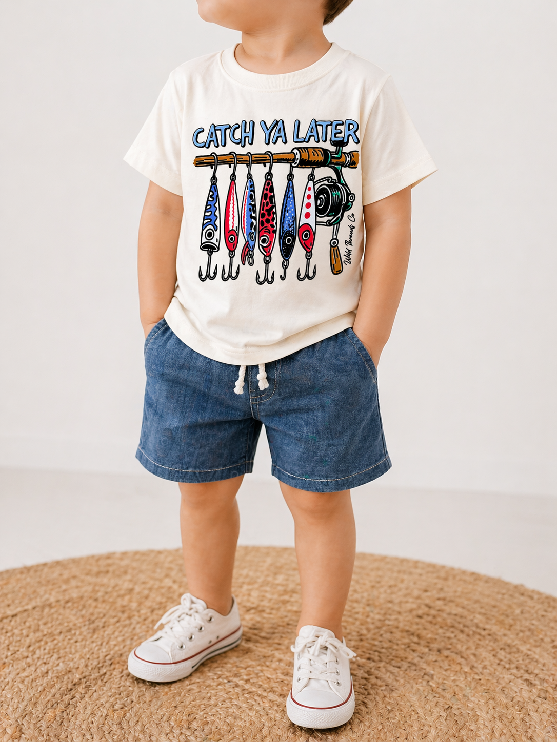 A young child stands on a round rug wearing the Wild Threads Co Catch Ya Later T-Shirt with colorful fishing lures, denim shorts, and white sneakers, hands tucked in their pockets.