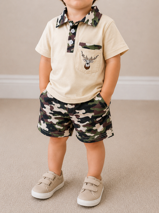 A young child indoors wears the Tiny Tot Bundles Camo Creek Polo Set: a beige polo with deer patch and camo collar, paired with camo shorts and beige sneakers, hands in pockets.