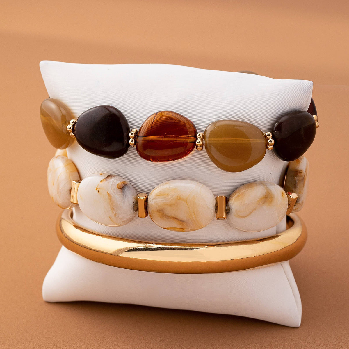The Boho Gold Bracelet Set by JOSSLYN by wall to wall features three bracelets displayed on a white cushion: one adorned with brown and gold stones, another with white marbled stones, and a shiny gold bangle—all set against a tan background.