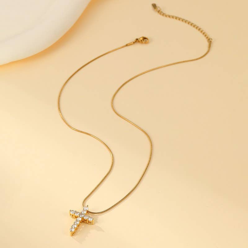 The Tarnish Free Cross Necklace by JOSSLYN by wall to wall, featuring a cross pendant embellished with small clear gemstones, is displayed on a cream-colored background.