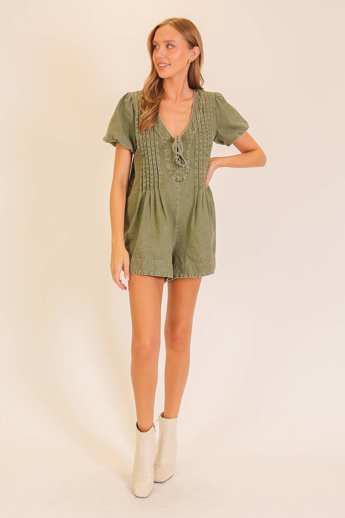 A woman stands against a plain background wearing the Lumiere Desert Muse Utility Romper in olive green, featuring short sleeves and a lace-up front detail, paired with white ankle boots.