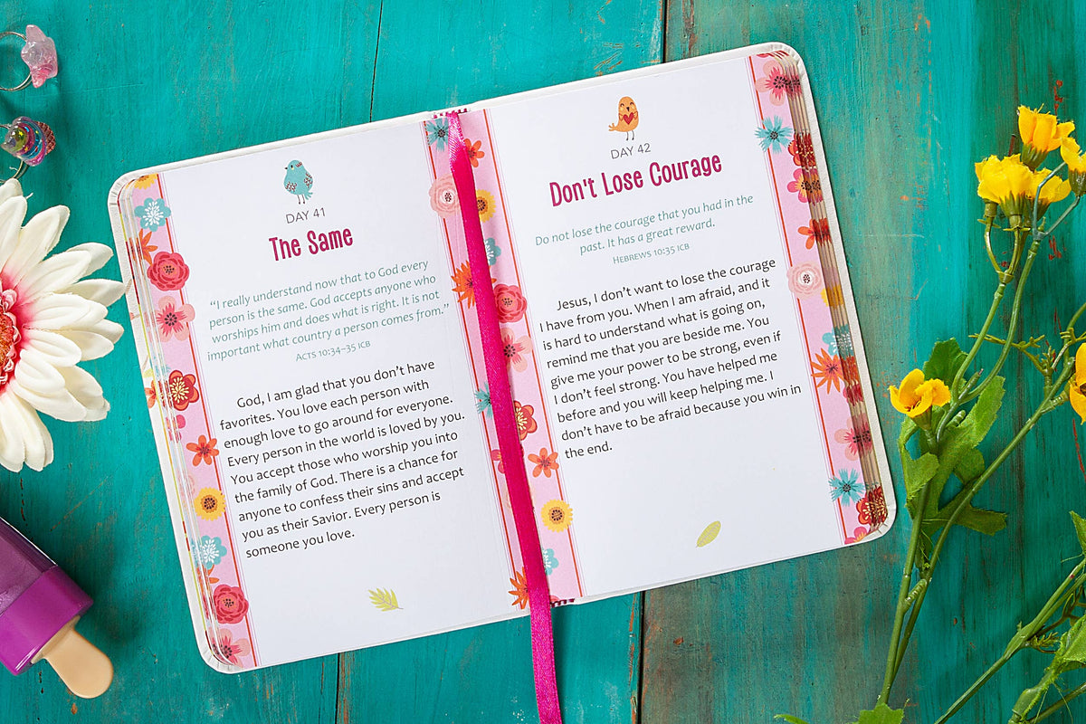 The 2-Minute Prayers for Girls Book by BroadStreet Publishing Group, LLC is open to days 41 and 42, displaying colorful floral borders and a pink ribbon bookmark. It rests on a turquoise wooden surface surrounded by scattered flower decorations.
