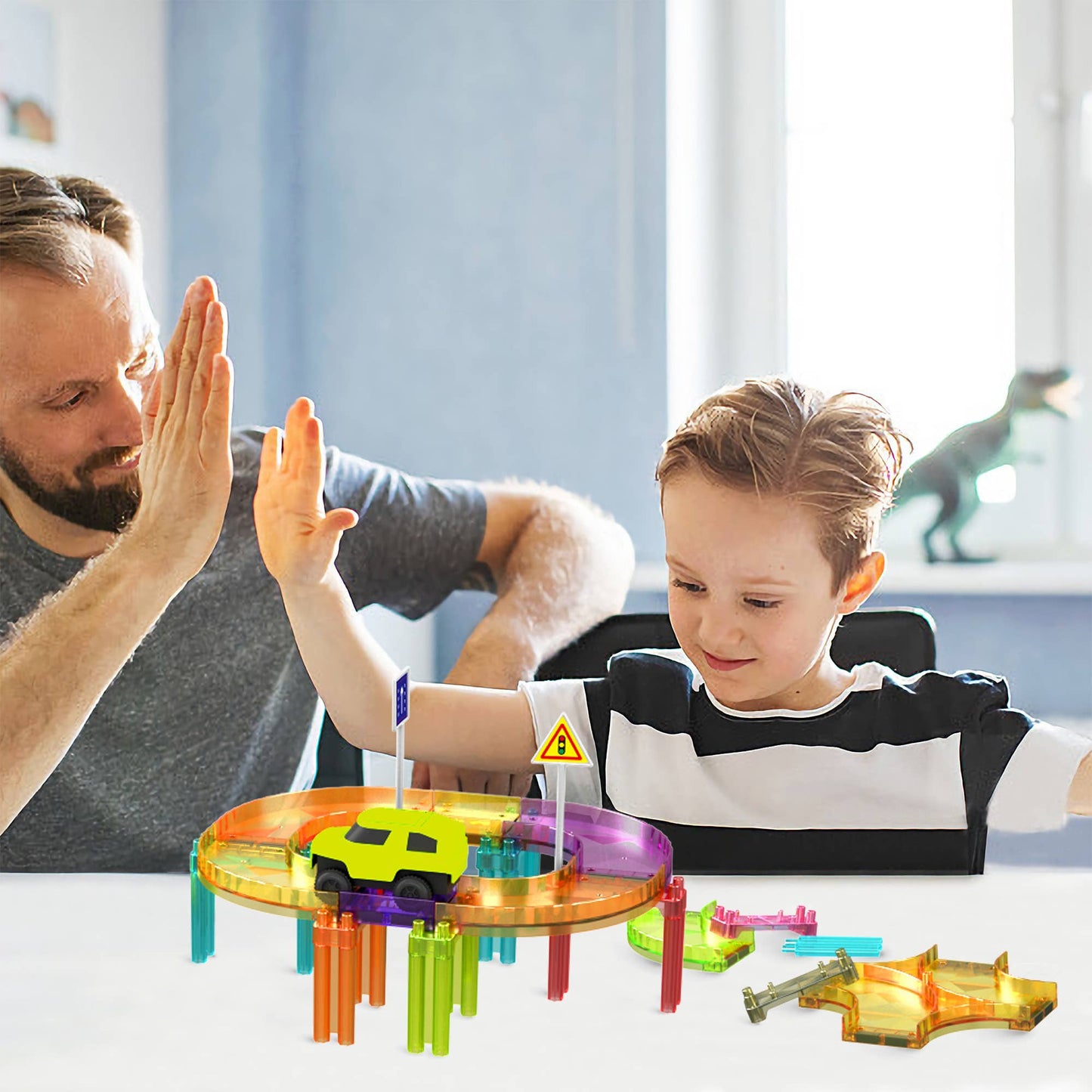 A smiling man and young boy high-five while playing with the PicassoTiles 40pc Travel Size Magnetic Tile Race Track Set by PicassoTiles in a blue room with a window and dinosaur figurine on the windowsill.